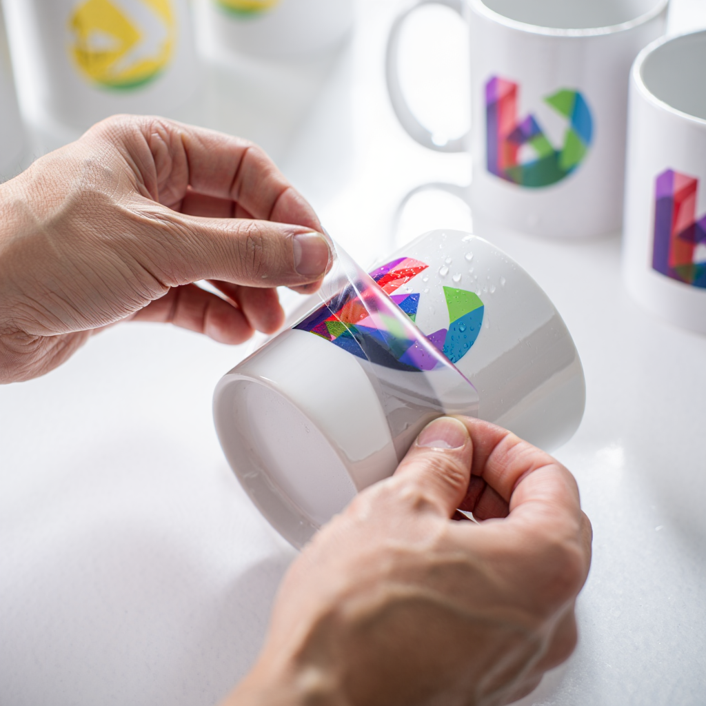 Person applying a colorful logo to a white mug with additional mugs in the background.